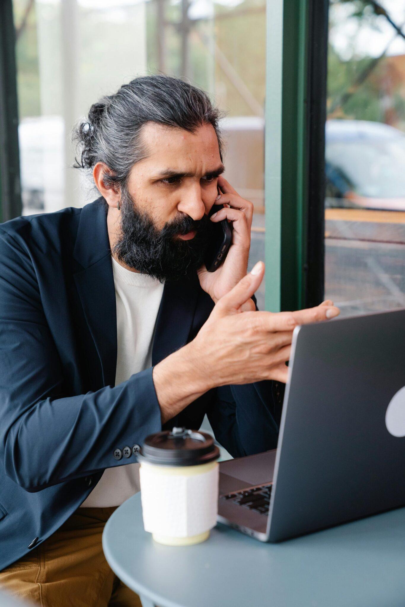 Bearded businessman multitasking with phone call and laptop in modern cafe setting.