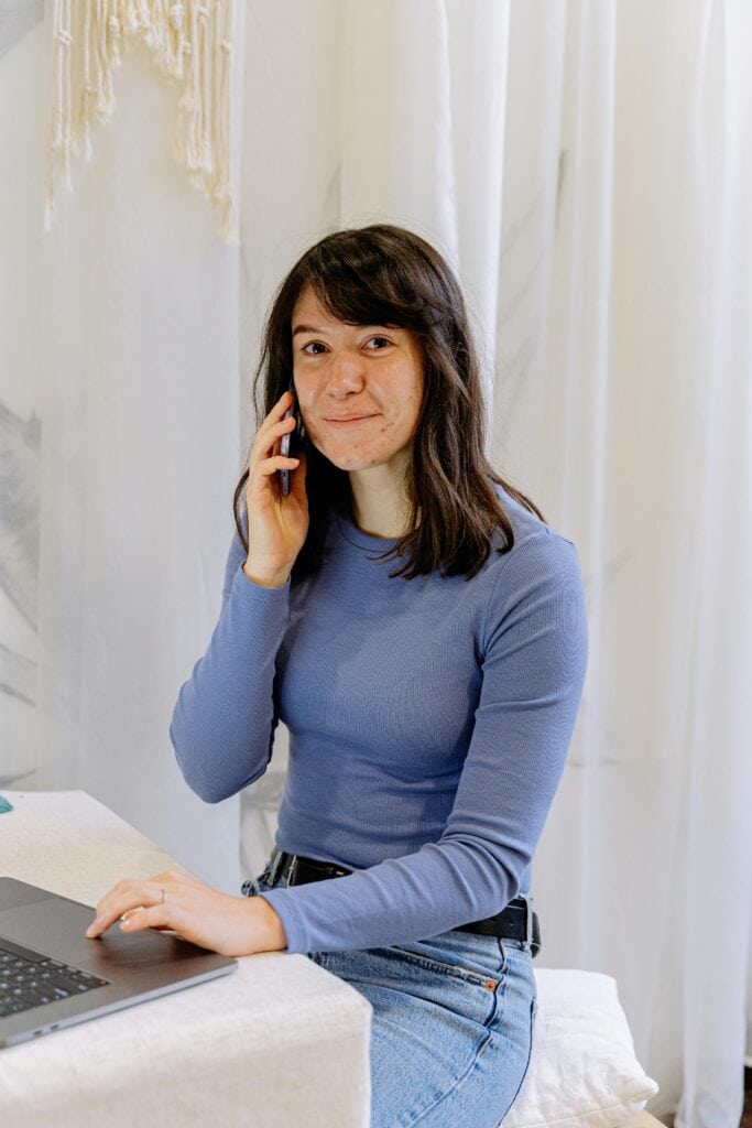 Contact 2 Smiling young woman talking on smartphone while working on laptop at home.