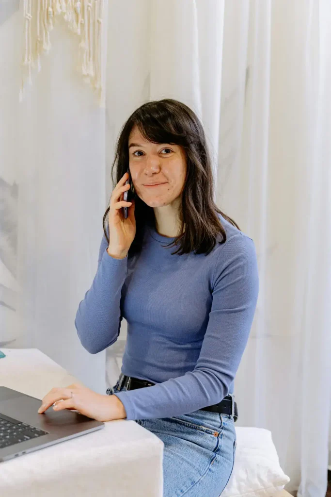 Smiling young woman talking on smartphone while working on laptop at home.