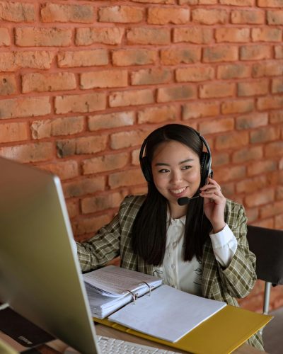 Home 11 Smiling Asian woman with headset working at computer in office with brick wall background.